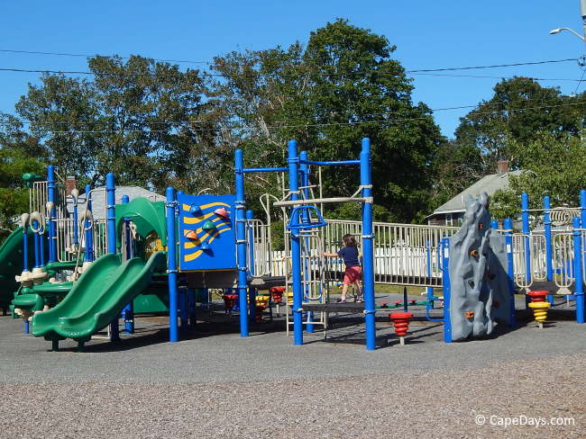 Colorful play structures at the All Access Playground in West Dennis, with slides, climbing equipment, and a child playing.