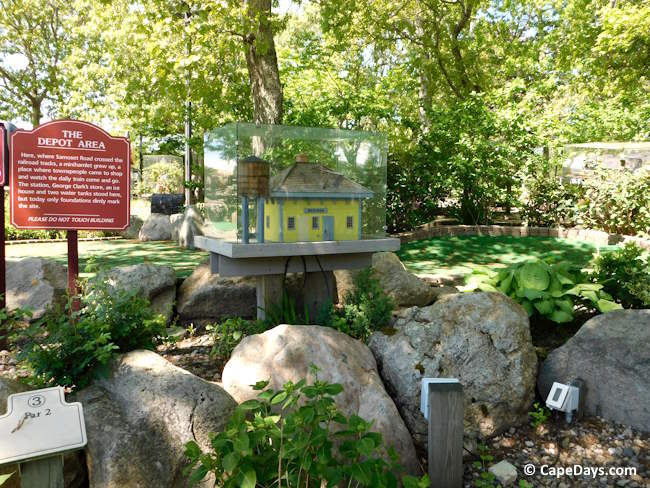 Miniature replica of Eastham’s old Samoset Road depot at Hole 3 of Arnold’s Mini Golf, with shrubs, boulders, and filtered sunlight through trees
