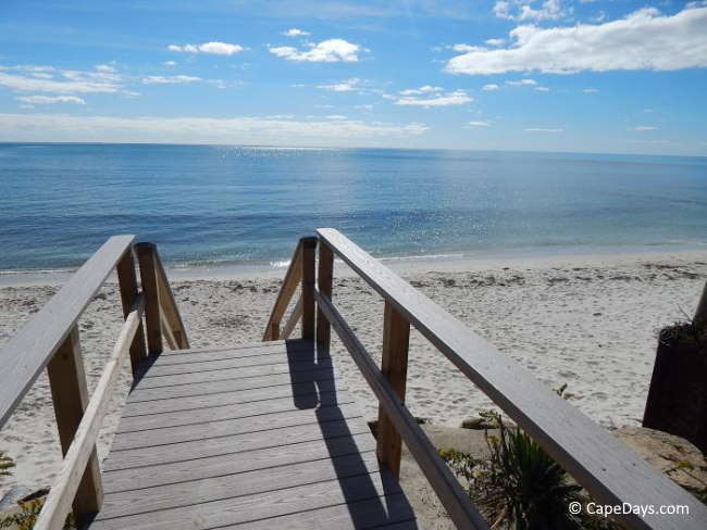 Wooden stairway leading to a narrow beach, bright blue sky with a few billowy white clouds, calm blue ocean