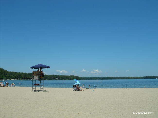 Blue sky over a pretty lake, people on the beach, lifeguard seated in umbrella-shaded tower