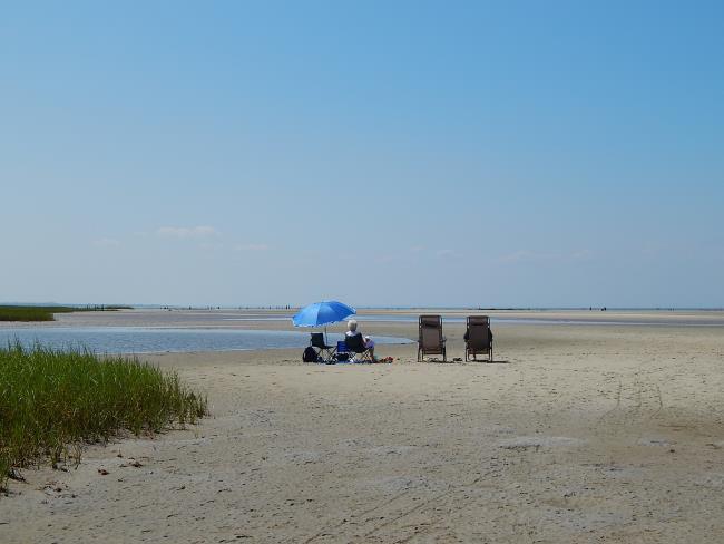 People relaxing in beach chairs, under a blue umbrella, during low tide at Rock Harbor