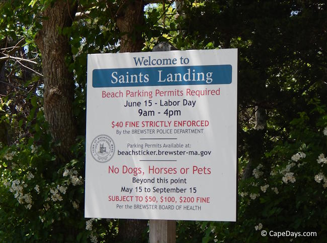 Permit-required sign at the entrance to a Cape Cod town beach, posted on a wooden stake with leafy trees behind it
