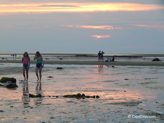 Two young ladies walking on the Brewster tidal flats at low tide near sunset, with reflections on the wet sand and other beachgoers in the distance.