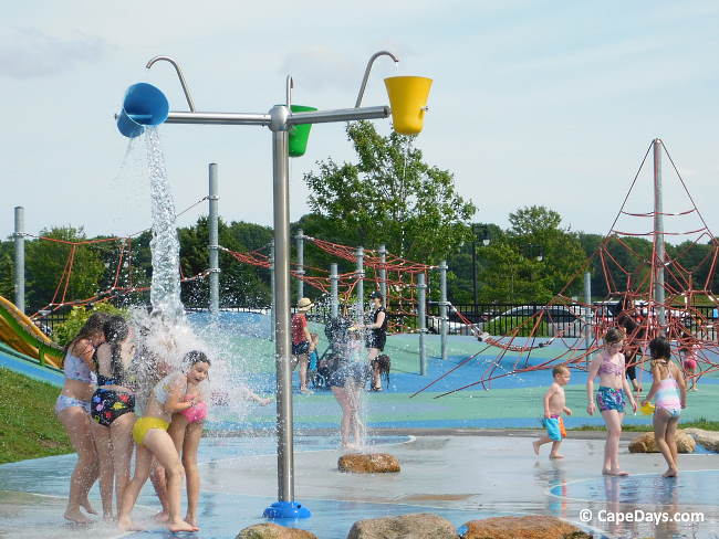 Kids standing under buckets pouring water from above at the Buzzards Bay splash pad near the Cape Cod Canal