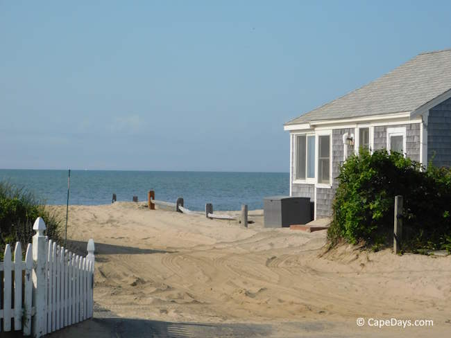 Weathered-shingle Cape Cod beach cottage with a white picket fence and ocean view on a sunny day