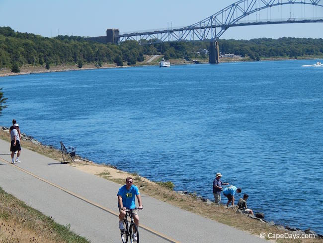 People sightseeing and bicycling along the Cape Cod Canal path on a wide section of the waterway.