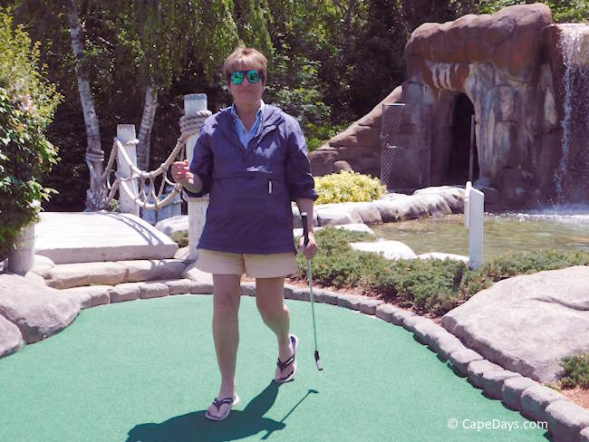 Person walking off the green with a putter, smiling, with a bridge, trees, and a waterfall cave exit in the background at a Cape Cod mini golf course