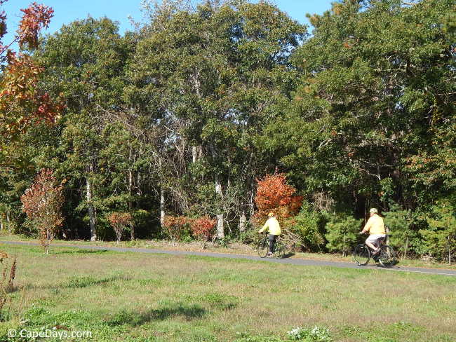 Two people riding bicycles on the Cape Cod Rail Trail in Dennis, with trees and open grassy area alongside the paved path.