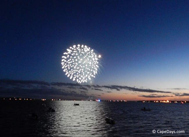 Firework exploding over the water at night, with boats and shoreline lights in the distance.