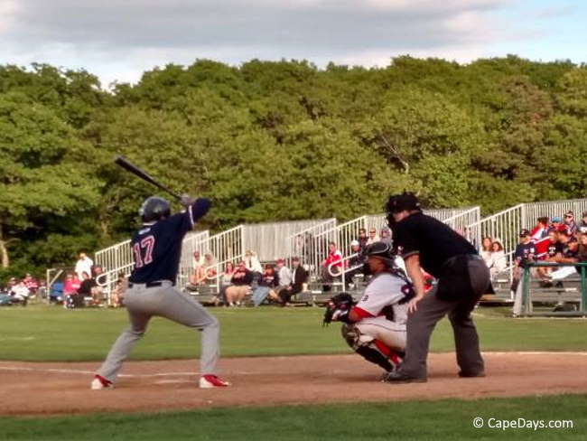 Cape Cod Baseball League player ready to swing his bat, with catcher and umpire behind the plate and spectators in the bleachers