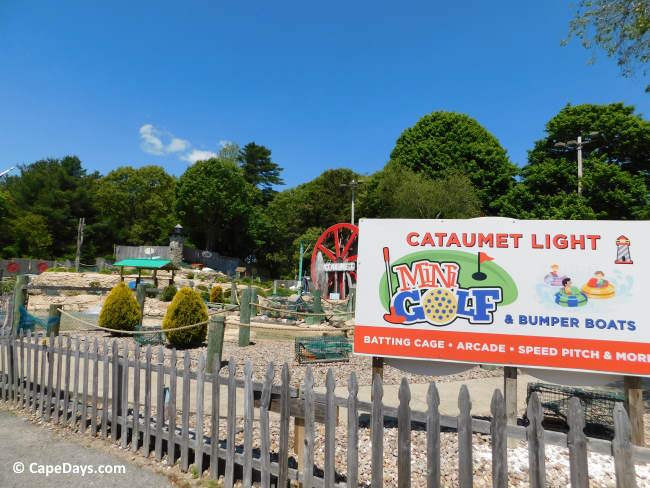 Sign for Cataumet Light Mini Golf with cartoonish art, weathered fence, and red water wheel in background.