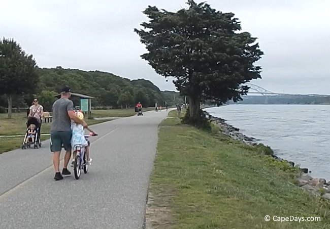 People walking, biking, and pushing strollers along the Cape Cod Canal path, with the Sagamore Bridge visible in the distance.