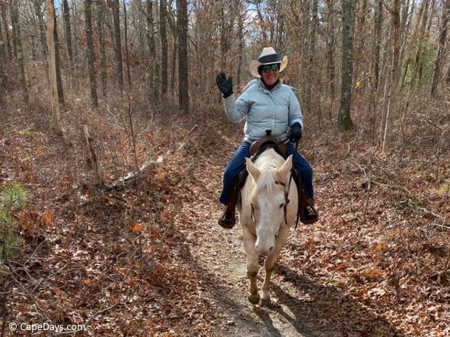 Woman wearing chilly-weather clothing and a western hat, riding a handsome palomino horse; bare trees and fallen leaves along the trail