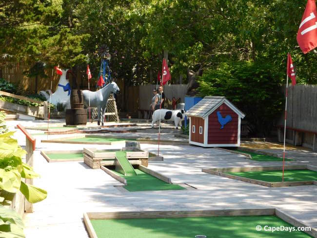 Farm-themed mini golf course at Gift Barn in Eastham, showing horse and cow statues, a red chicken coop, hole flags, and trees and picket fence lining the back of the course.