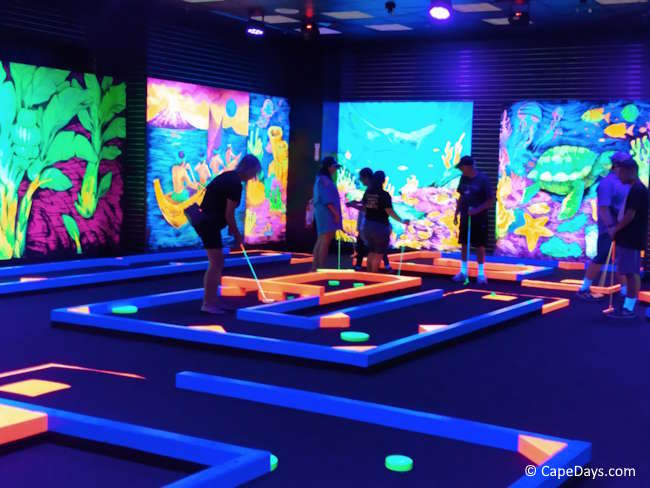 Glow-in-the-dark mini golf course at Cape Cod Mall in Hyannis, with black lights, colorful murals, and players on several holes.