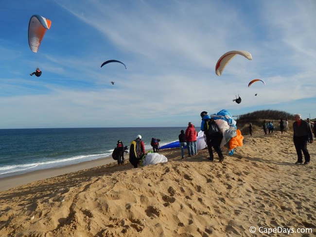People standing on top of dune watching hang gliders over the beach