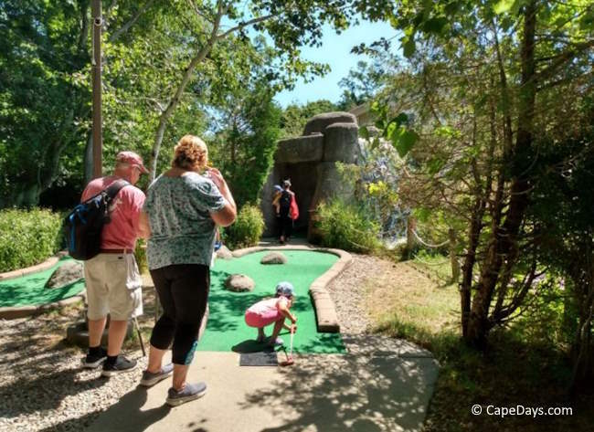 Toddler crouching on putting green at Harbor Lights Mini Golf, reaching for golf ball, as adults look on.