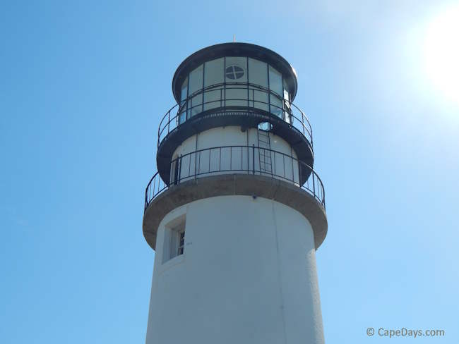 View looking up the side of Highland Light from ground level, with the lighthouse tower stretching toward a bright blue sky.