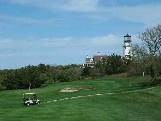 View of a golf cart on the green at Highland Links Golf Course. The white tower of Cape Cod Lighthouse and the weathered-shingled keeper's house in the background