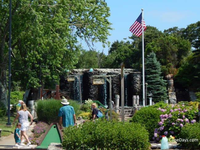 Players on putting green at Holiday Hill Mini Golf, with hydrangea bush, waterfall, and American flag nearby