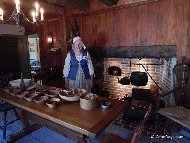 Interior of the Josiah Dennis Manse in Dennis, showing a colonial-era kitchen with a hearth, wooden table, and a docent in period costume.