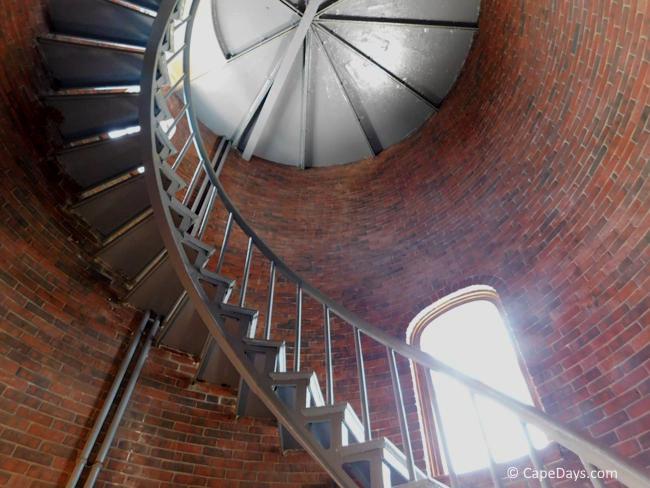 Winding metal staircase inside a brick lighthouse tower with light showing through a window