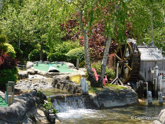 Waterwheel, waterfall, red Adirondack chair, and putting green under mature trees at Lightning Falls Mini Golf.