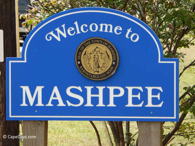 Blue road sign with gold medallion of the town of Mashpee and white print that says "Welcome to Mashpee", gold