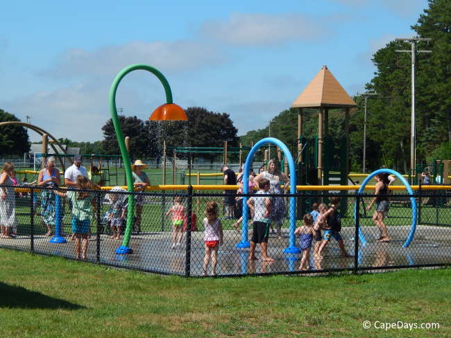 Fenced-in splash pad with colorful water features, kids playing while parents watch
