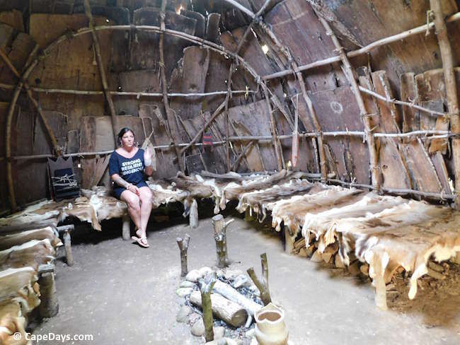 Interior of a Wampanoag wetu at the Mashpee Wampanoag Museum, showing hide-covered sleeping platforms, a fire pit, and the curved wood frame.