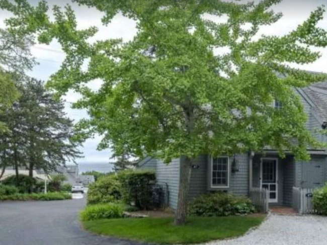 Gray-shingled condo cottage, mature trees and green lawn, view of ocean in background