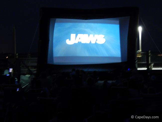 Dark sky surrounding a large outdoor movie screen showing the intro for the film "Jaws"