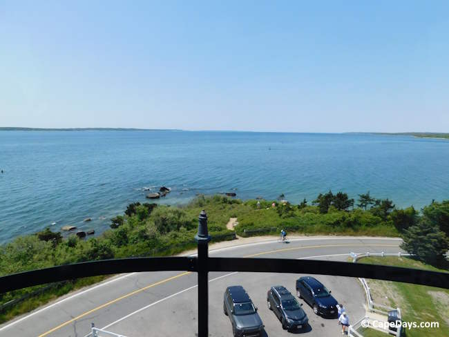 View from the lantern room at Nobska Light, showing the black railing, Vineyard Sound, a glimpse of Martha’s Vineyard, and the parking lot and road below.
