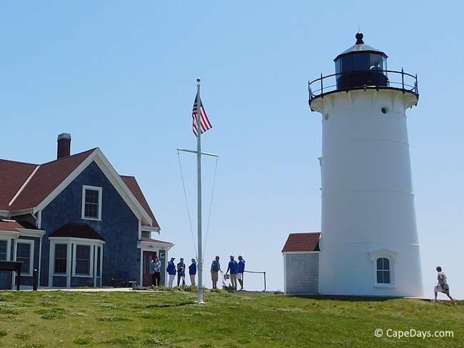 View of Nobska Light and the Keeper’s House from the parking lot, with a group of visitors and tour guides gathered near the lighthouse on a sunny morning.