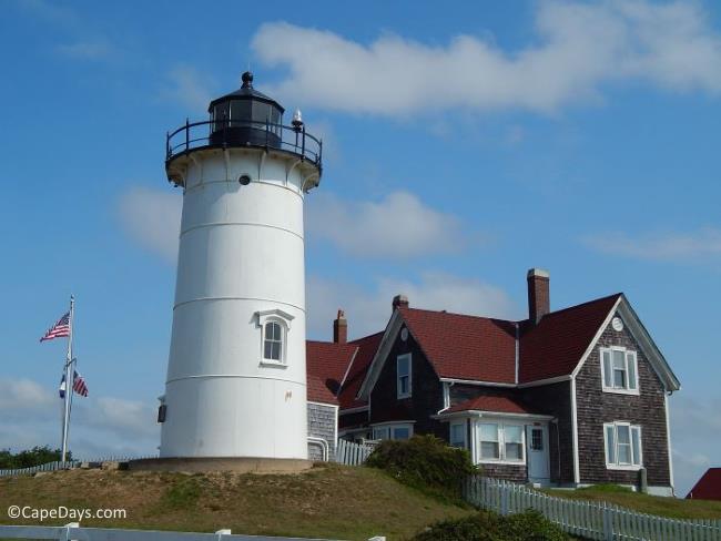 Weathered shingled keepers house alongside white lighthouse tower, flag pole and blue sky in the background