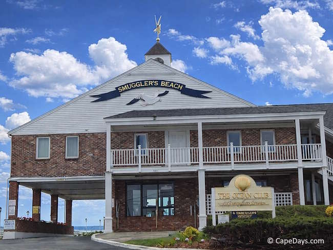Brick-and-white lodging building with balconies, a Smuggler’s Beach sign, and ocean visible beyond in South Yarmouth on Cape Cod.