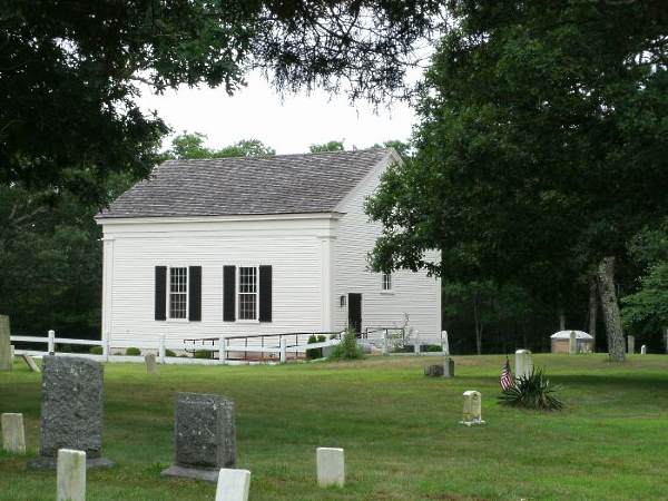 White building with black shutters, burial grounds with green grass, large trees and old headstones
