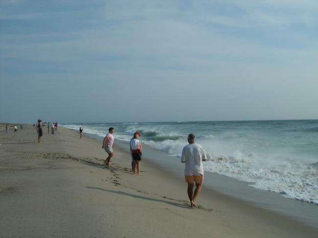 Sun breaks through high clouds,  beachgoers stand near the water's edge watching post-storm waves crashing on the beach.
