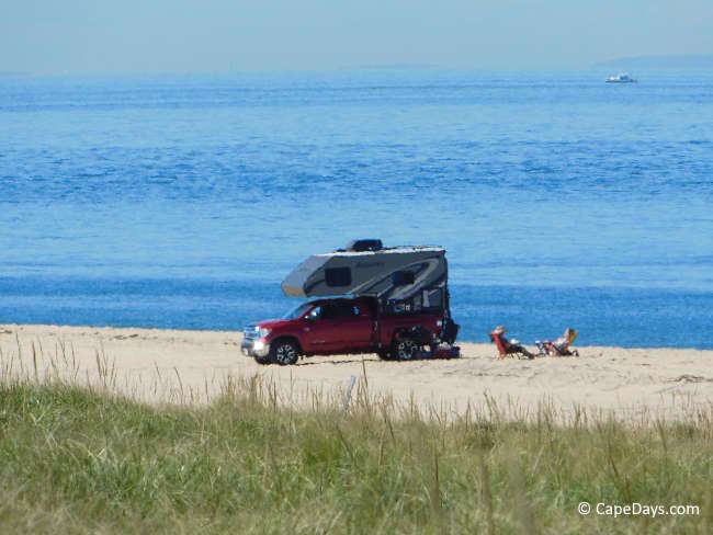 Self-contained vehicle parked on a Cape Cod beach with two people relaxing in beach chairs, enjoying ocean views on a sunny day