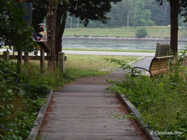 Pathway from Bourne Inclusive Playground to the Cape Cod Canal, with a partial wooden walkway, a bench, and trees framing the view of the water Pathway from Bourne Inclusive Playground to the Cape Cod Canal, with a partial wooden walkway, a bench, and trees framing the view of the water
