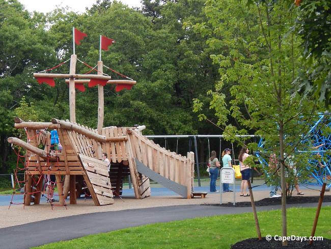 Large pirate ship-themed play structure in the older kids’ area at Sandy Pond Playground in Yarmouth, with orange flags on two masts, a child climbing, and adults standing nearby Large pirate ship-themed play structure in the older kids’ area at Sandy Pond Playground in Yarmouth, with orange flags on two masts, a child climbing, and adults standing nearby