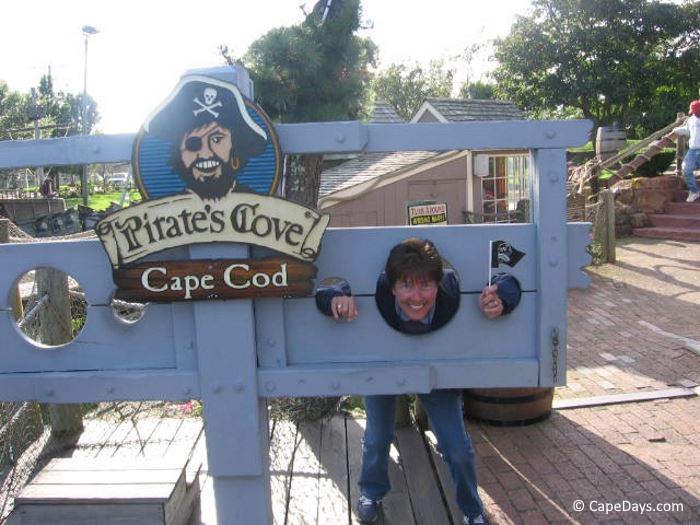 Woman posing in wooden stocks at Pirate’s Cove Mini Golf, holding small pirate flag, with themed signage nearby