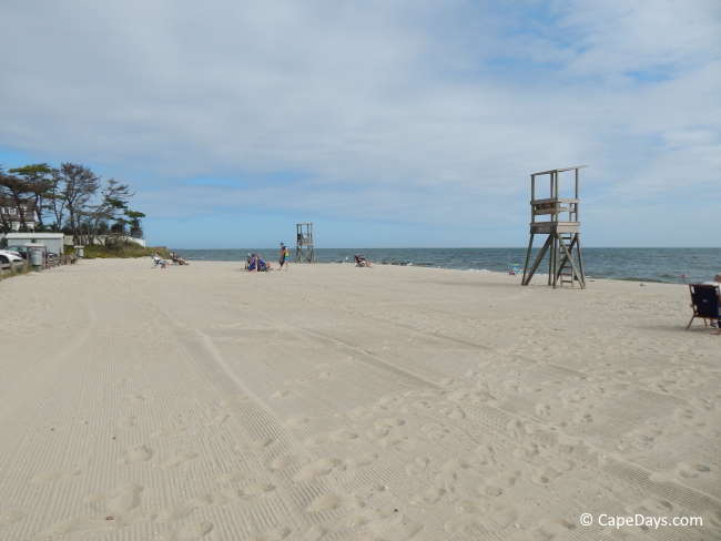 Puffy clouds in a blue sky over a dark blue ocean, wooden lifeguard stations on the beach, beachgoers in the distance