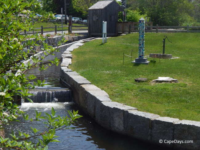 Water flowing down the herring run steps near Dexter’s Grist Mill in Sandwich, with ticket booth, lawn, hand-pump, and colorful sculpture in view Water flowing down the herring run steps near Dexter’s Grist Mill in Sandwich, with ticket booth, lawn, hand-pump, and colorful sculpture in view