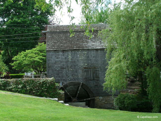 Historic Dexter Grist Mill building with weathered shingles, water wheel, surrounded by green trees and lawn Historic Dexter Grist Mill building with weathered shingles, water wheel, surrounded by green trees and lawn