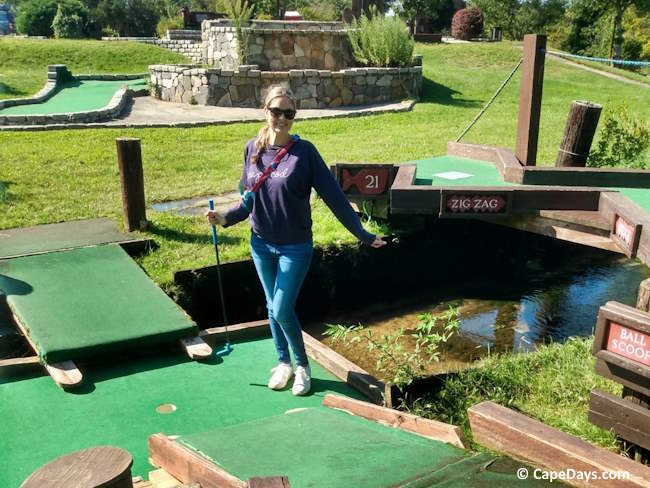 Young woman celebrating a hole-in-one at Sandwich Mini Golf, standing on a putting green with arms outstretched. Several greens, natural grass, and part of a water feature are also visible. 