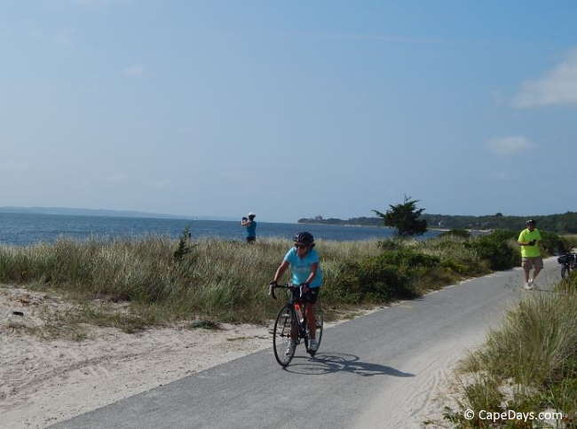 A cyclist riding along the Shining Sea Bikeway in Falmouth, with one person walking and another stopped alongside the trail taking a photo of Vineyard Sound.