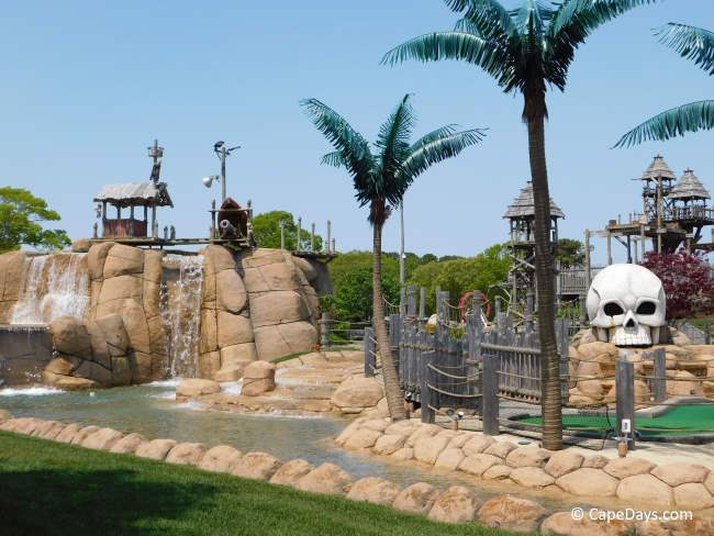 Large skull statue wearing a red, white, and blue headband next to a waterfall at Skull Island Mini Golf, with palm trees and tiki huts in the background.
