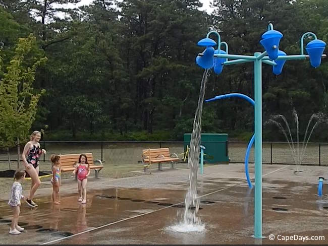 Children playing on the splash deck at Sandy Pond Recreation Area in Yarmouth, Cape Cod, with water splashing from blue overhead buckets and ground-level sprays in the background Children playing on the splash deck at Sandy Pond Recreation Area in Yarmouth, Cape Cod, with water splashing from blue overhead buckets and ground-level sprays in the background