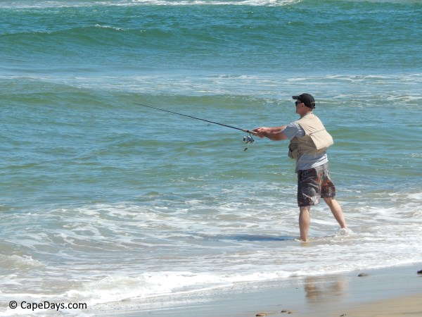 Man wearing a tan shirt, brown cargo shorts, a ball cap and a fishing backpack, casting his line into the Atlantic Ocean surf Man wearing a tan shirt, brown cargo shorts, a ball cap and a fishing backpack, casting his line into the Atlantic Ocean surf